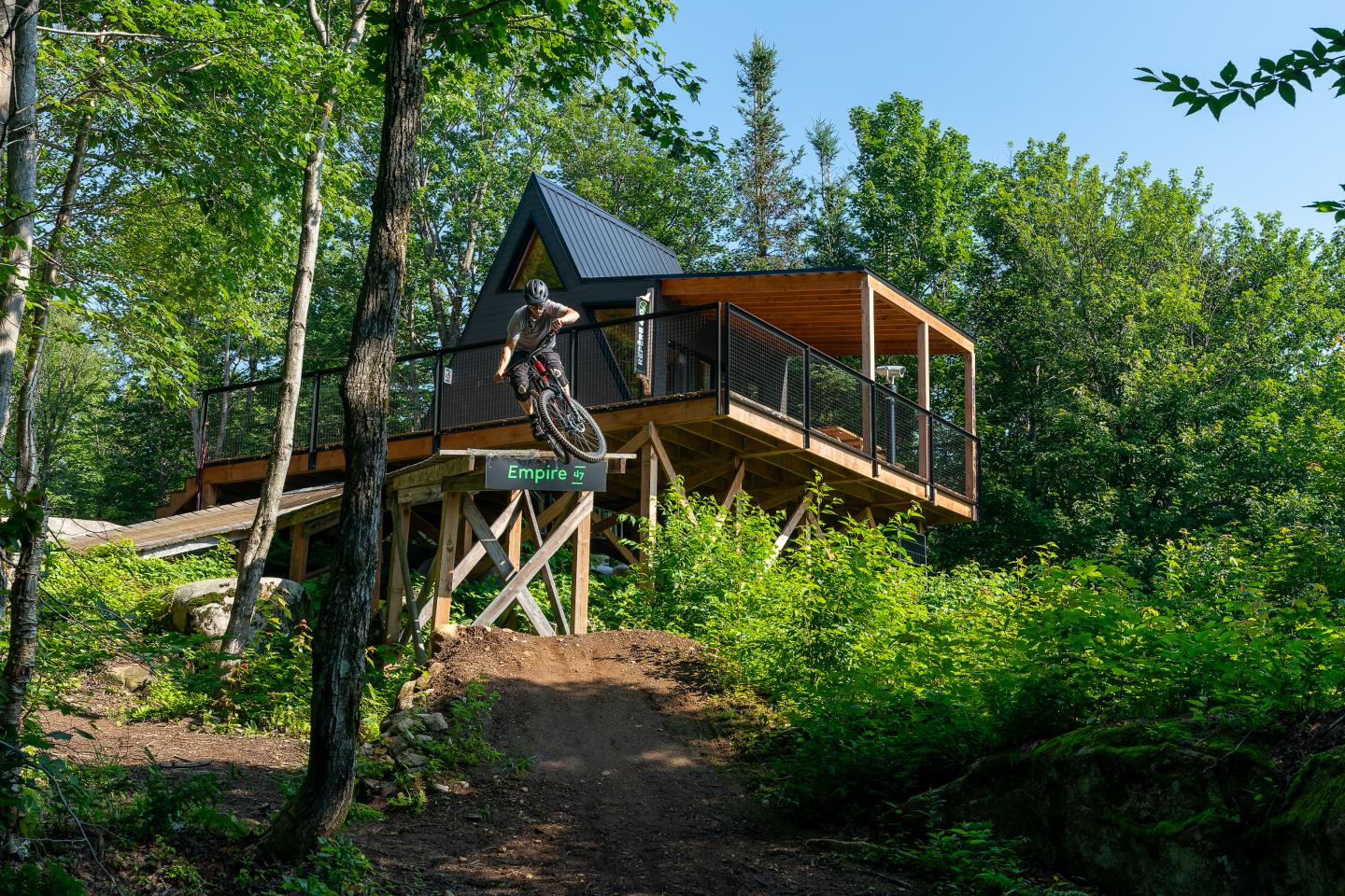 Un cycliste s'élance d'une drop, avec un chalet d'accueil moderne en arrière-plan.