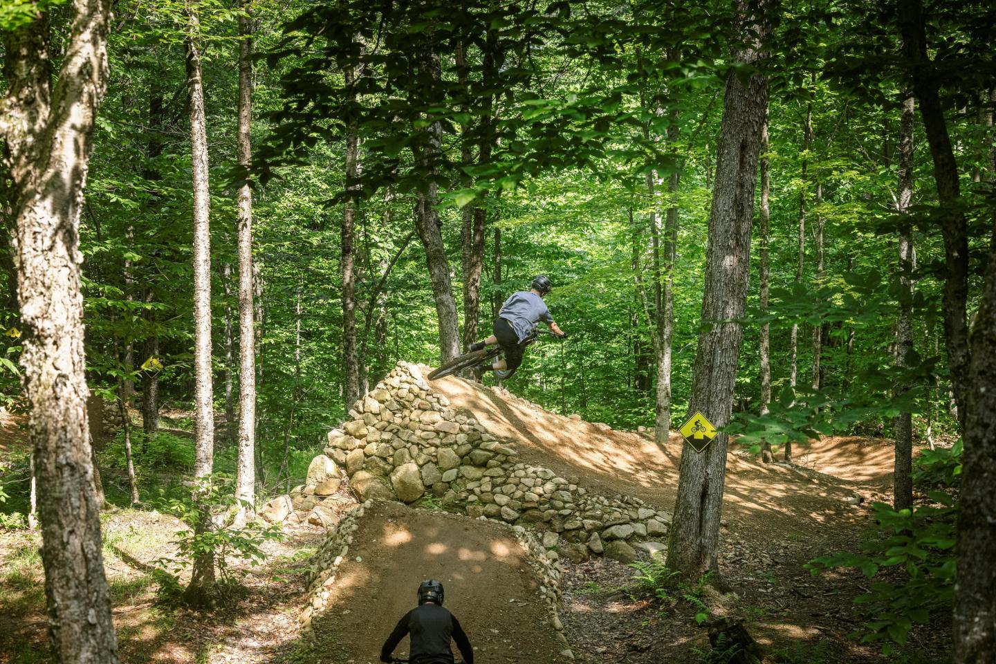 Deux cyclistes sur un sentier en forêt, l'un sautant une rampe.