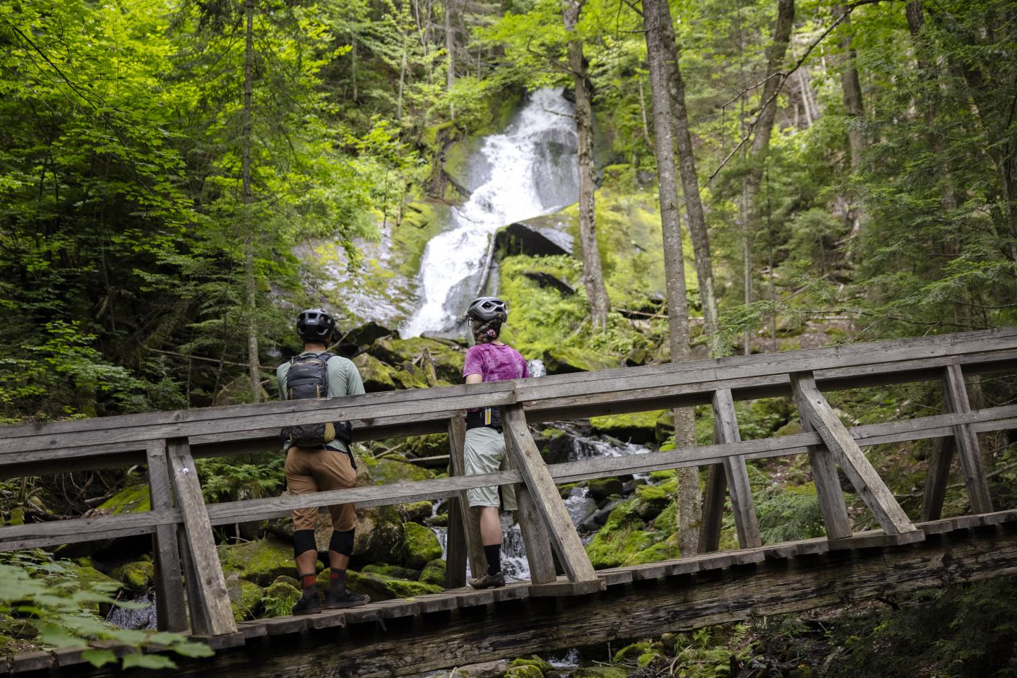 Trois personnes sur un pont en bois face à une cascade en forêt.