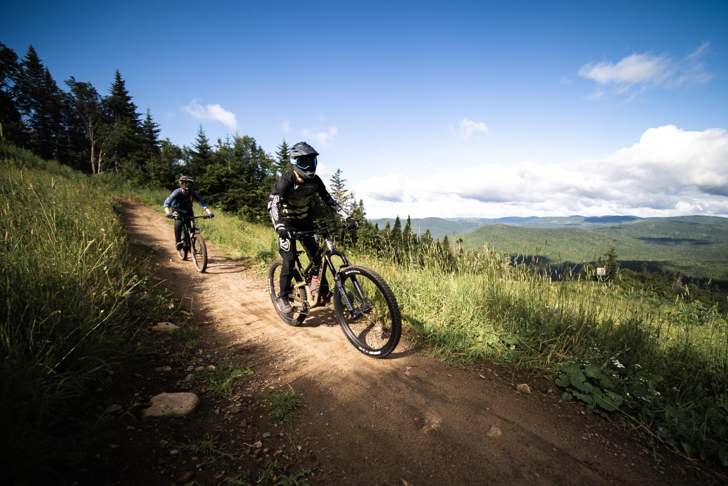 Cyclistes en montagne sur un chemin en terre, ciel bleu et nuages.